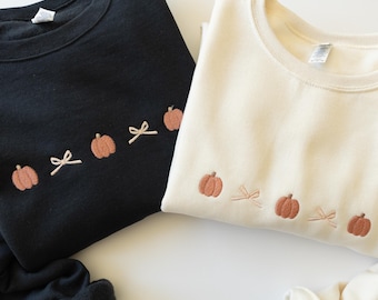 Puede incluir: Una sudadera negra y una sudadera color crema, cada una con una fila horizontal bordada de pequeñas calabazas naranjas alternadas con lazos de color beige claro en el área del pecho. Ambas prendas tienen mangas largas y cuello redondo.