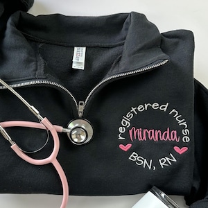 May include: Black 1/4 zip sweatshirt with a white embroidered design that says "registered nurse Miranda BSN, RN" with two pink hearts. A pink stethoscope and a white travel mug are also in the image.