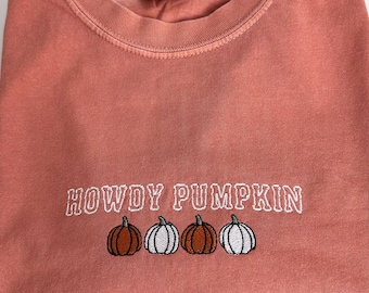 Puede incluir: Una sudadera de cuello redondo color terracota con texto bordado en blanco que dice HOWDY PUMPKIN. Debajo del texto hay cuatro pequeñas calabazas bordadas en colores alternos naranja y blanco con tallos y contornos verde oscuro.