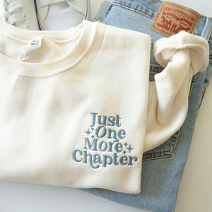 Puede incluir: Una sudadera color crema con el texto bordado "Just One More Chapter" en azul, combinada con vaqueros de mezclilla de lavado claro y zapatillas blancas. Los vaqueros tienen una etiqueta de Levi Strauss & Co.