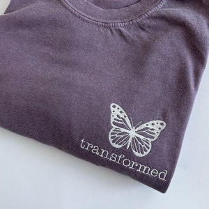 Può includere: Una maglietta viola con una farfalla bianca e la parola "trasformato" ricamata sul davanti.