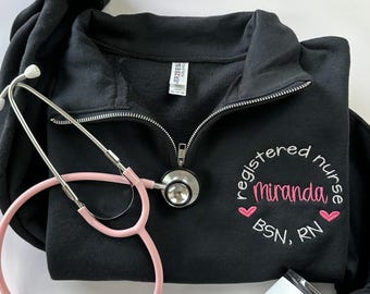 May include: Black 1/4 zip sweatshirt with a white embroidered design that says "registered nurse Miranda BSN, RN" with two pink hearts. A pink stethoscope and a white travel mug are also in the image.