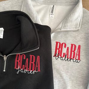 Puede incluir: Dos sudaderas con cremallera de cuarto de c&iacute;rculo, una negra y otra gris. Ambas presentan texto bordado en rojo: "BCABA" sobre un nombre en cursiva. La sudadera negra tiene "Javier" bordado en blanco. La gris tiene "Valeria" bordado en negro.