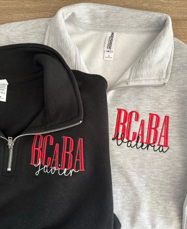 Puede incluir: Dos sudaderas con cremallera de cuarto de c&iacute;rculo, una negra y otra gris. Ambas presentan texto bordado en rojo: "BCABA" sobre un nombre en cursiva. La sudadera negra tiene "Javier" bordado en blanco. La gris tiene "Valeria" bordado en negro.
