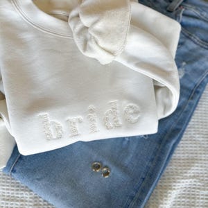 Puede incluir: Sudadera blanca con la palabra "bride" bordada en blanco en la parte delantera. La sudadera está doblada y encima de un par de pantalones vaqueros azules.