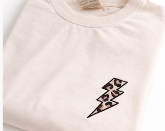 Może przedstawiać: Kremowy t-shirt z okrągłym dekoltem, ozdobiony małym haftowanym błyskawicą na lewej piersi. Błyskawica ma czarny kontur i jest wypełniona beżowo-czarnym wzorem w panterkę. Przy kołnierzu widoczna jest metka z instrukcją prania.