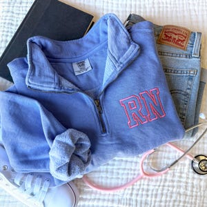 May include: A light blue fleece quarter-zip sweatshirt with the letters "RN" embroidered in pink on the front. The sweatshirt is laid out on a white surface with a stethoscope and a pair of jeans.