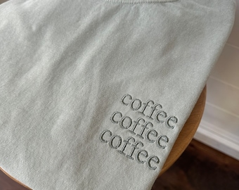 May include: A light sage green crewneck sweatshirt featuring the word coffee embroidered three times in a stacked vertical arrangement on the lower right side in a dark grey thread. The garment has a ribbed collar and a soft, washed texture.