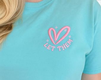 Puede incluir: Una camiseta azul claro con un diseño bordado en el pecho. El bordado consiste en el contorno de un corazón rosa, pequeños detalles de estrellas rosas y el texto rosa "LET THEM" colocado en un arco debajo de la forma del corazón.