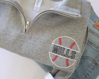 May include: A heather gray quarter-zip sweatshirt featuring an embroidered baseball graphic with red stitching and the name MILLS in black block letters. The sweatshirt has a silver zipper and is layered over a pair of light blue denim jeans.