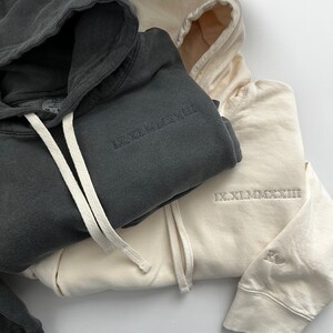 Puede incluir: Dos sudaderas con capucha, una en gris oscuro y otra en crema, con la inscripci&oacute;n en n&uacute;meros romanos "IX.XI.MMXXIII" en cada una.