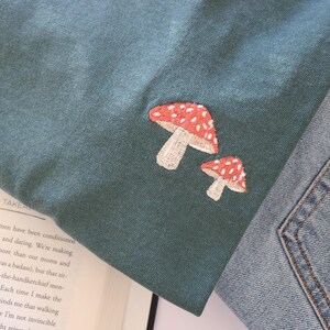 Pu&ograve; includere: Una maglietta blu turchese con un designo di fungo ricamato in rosso e bianco. La maglietta &egrave; piegata e appoggiata su un libro.
