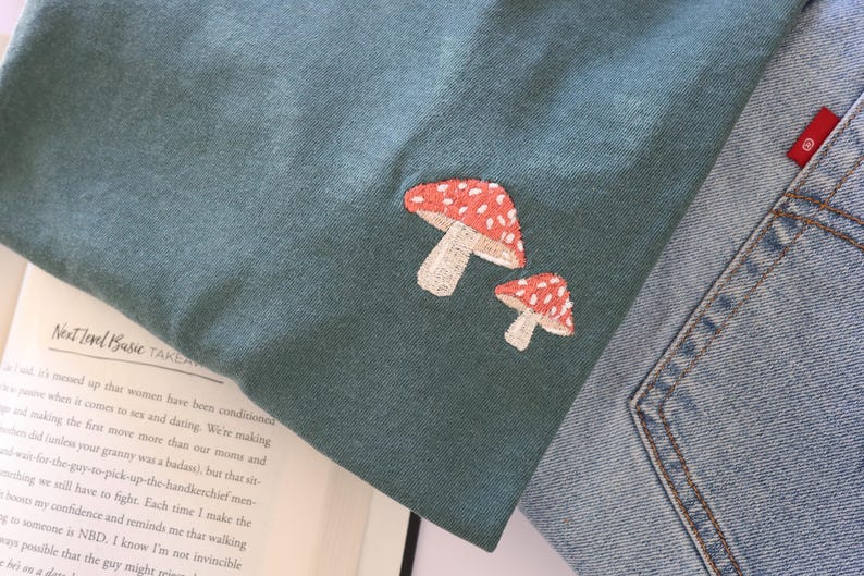 Pu&ograve; includere: Una maglietta blu turchese con un designo di fungo ricamato in rosso e bianco. La maglietta &egrave; piegata e appoggiata su un libro.