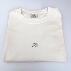 Może przedstawiać: Kremowy t-shirt z okrągłym dekoltem i wyhaftowanymi zielonymi literami JRH na środku klatki piersiowej. Wewnętrzna metka przy kołnierzu zawiera nazwę marki Comfort Colors oraz rozmiar XL-TG-EG. Odzież jest starannie złożona na płaskiej powierzchni.