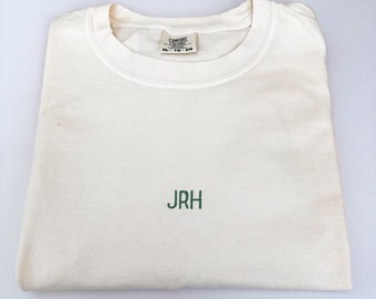 Puede incluir: Una camiseta de cuello redondo color blanco hueso con las letras JRH bordadas en verde en el centro del pecho. La etiqueta interior del cuello muestra la marca Comfort Colors y la talla XL-TG-EG. La prenda está doblada sobre una superficie.