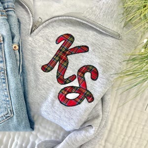 May include: A light gray sweatshirt with a quarter-zip neckline and the embroidered initials "KS" in a red, green, and yellow plaid pattern. Light blue denim jeans are visible on the left side of the image.