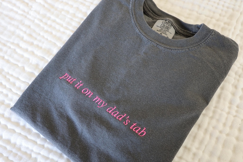 以下が含まれることがあります： ピンク色の刺繍で「put it on my dad's tab」と書かれたチャコールグレーのTシャツ。折りたたまれたシャツは、白くテクスチャ加工された表面に置かれています。クルーネックで、Comfort Colorsのタグが付いています。