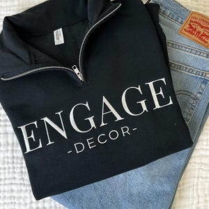 May include: A black quarter-zip sweatshirt featuring the white text "ENGAGE -DECOR-" on the front. The garment has a silver-toned metal zipper at the collar and a Jerzees brand label inside the neck. It is positioned next to light blue denim jeans.