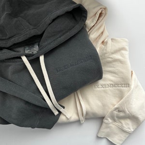 May include: Two hooded sweatshirts, one dark gray and one light beige. The gray sweatshirt has the text "IX.XI.MMXIII" embroidered on the front. The beige sweatshirt has the text "IX.XI.MMXIII" embroidered on the front and the text "Comfort Colors" on the tag.