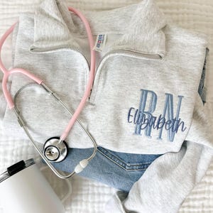 Könnte beinhalten: Hellgraues Sweatshirt mit Viertelreißverschluss und blauem, gesticktem Text "RN Elizabeth". Ein rosa Stethoskop und ein weißer Becher befinden sich ebenfalls im Bild und liegen auf einer Jeans.