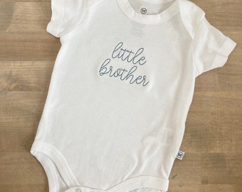 May include: White baby onesie with short sleeves and snaps at the bottom. The onesie has the text "little brother" embroidered in gray on the front.