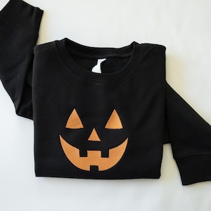Pode incluir: Uma camisola preta de manga comprida com um grande gráfico laranja centrado de uma cara de abóbora de Halloween. O design inclui dois olhos triangulares, um nariz triangular e um sorriso largo com dentes. Gola redonda e punhos canelados.