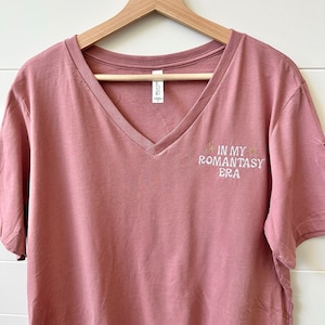 Puede incluir: Una camiseta de cuello en V rosa polvoriento con mangas cortas. La camiseta tiene el texto bordado "IN MY ROMANTASY ERA" en blanco con detalles dorados. La camiseta está colgada en una percha de madera.