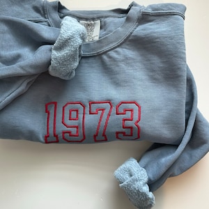 Puede incluir: Una sudadera azul grisáceo con el año "1973" bordado en rojo. Los puños están enrollados, revelando un forro azul más claro. La etiqueta dice "Comfort Colors".