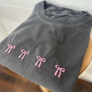 May include: A charcoal gray crew neck t-shirt featuring four small, embroidered light pink bows arranged in a horizontal row across the chest. The fabric has a slightly faded, washed-out appearance, and the shirt is folded neatly.