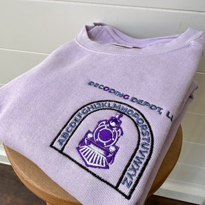 May include: A lavender sweatshirt with an embroidered design of a train engine in a circle with the letters of the alphabet around it. The text "Decoding Depot" is above the train engine.