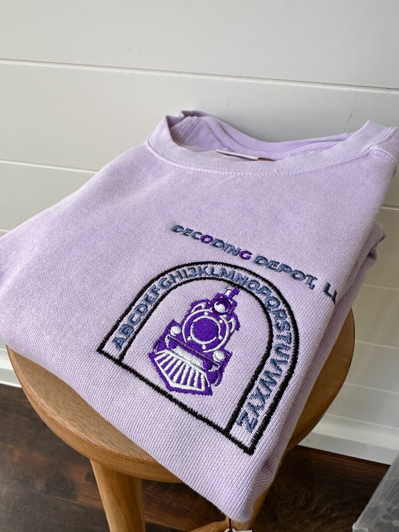 May include: A lavender sweatshirt with an embroidered design of a train engine in a circle with the letters of the alphabet around it. The text "Decoding Depot" is above the train engine.