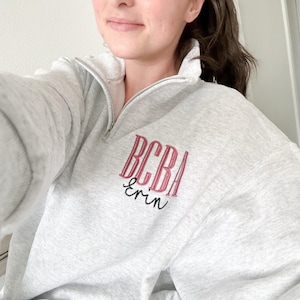 May include: A light gray pullover sweatshirt with a quarter-zip neckline. The sweatshirt features the embroidered letters "BCBA" in a burgundy color, with the name "erin" embroidered below in a cursive font. The sweatshirt is a casual style.