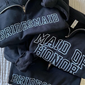 May include: Three black quarter-zip sweatshirts featuring embroidered text in light blue thread. One sweatshirt displays the word BRIDESMAID, another reads MAID OF HONOR, and the third features BRIDESMAID. Each garment has a silver-toned zipper.