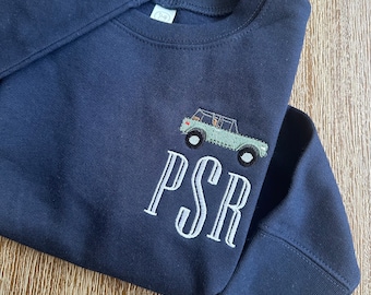 May include: A navy blue long-sleeve sweatshirt featuring an embroidered light green off-road vehicle above the letters PSR in a white serif font. The garment has a ribbed crew neckline and ribbed cuffs, with a small white tag inside the collar.