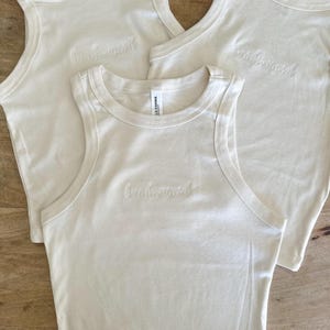 Pu&ograve; includere: Tre canotte bianco sporco con la parola "bridesmaid" ricamata sul davanti. I top sono realizzati in un materiale morbido e hanno uno stile classico e casual. Le canotte sono su una superficie di legno.