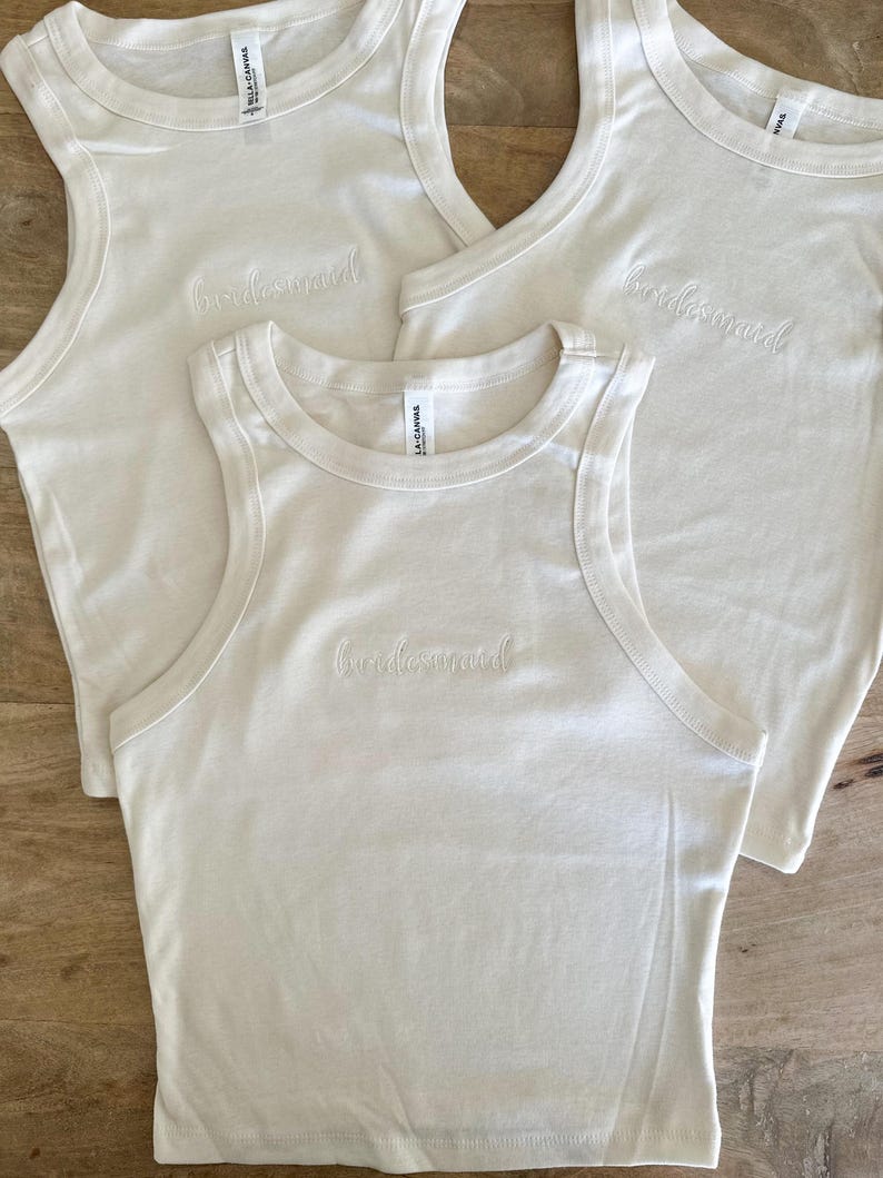 Pu&ograve; includere: Tre canotte bianco sporco con la parola "bridesmaid" ricamata sul davanti. I top sono realizzati in un materiale morbido e hanno uno stile classico e casual. Le canotte sono su una superficie di legno.