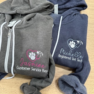 May include: Three hooded zip-up sweatshirts in shades of gray and blue. One gray sweatshirt features the name "Jasmine" and "Customer Service Rep" embroidered in pink. The other blue sweatshirt has "Michelle Registered Vet Tech" embroidered in white.