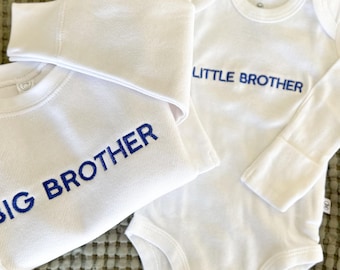 May include: White baby clothes set with blue embroidered text. The set includes a long-sleeved onesie with "LITTLE BROTHER" and a long-sleeved shirt with "BIG BROTHER". A matching white beanie is also included. The clothes are made of soft material.