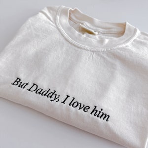 May include: An off-white crew neck t-shirt featuring the black embroidered text "But Daddy, I love him" across the front. The garment has a ribbed collar and is folded to display the neckline and the embroidered phrase clearly on the fabric.