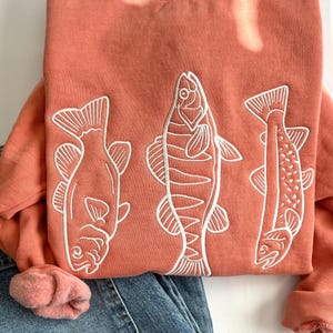 May include: A coral-coloured crewneck sweatshirt featuring three white embroidered fish designs on the front. The sweatshirt has long sleeves with ribbed cuffs and a ribbed neckline. The fabric has a soft, fleece-like texture throughout.