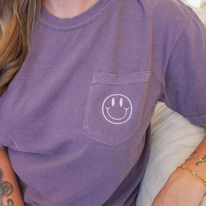 May include: A purple t-shirt with a white smiley face embroidered on the pocket.