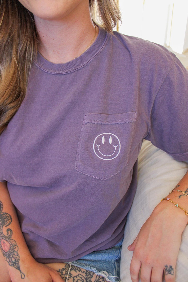May include: A purple t-shirt with a white smiley face embroidered on the pocket.