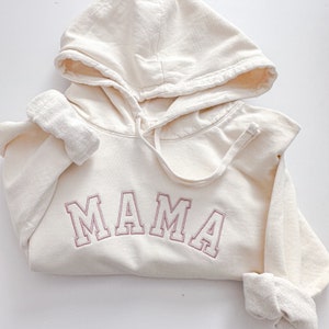 Könnte beinhalten: Ein cremefarbener Hoodie mit einer Kapuze mit Kordelzug und dem Wort "MAMA" in rosa auf der Vorderseite gestickt.