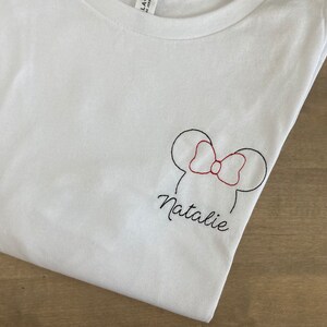 May include: White t-shirt with a black embroidered outline of Mickey Mouse ears with a red bow and the name Natalie.