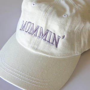 May include: A white baseball cap with the word "MOMMIN'" embroidered in purple on the front.