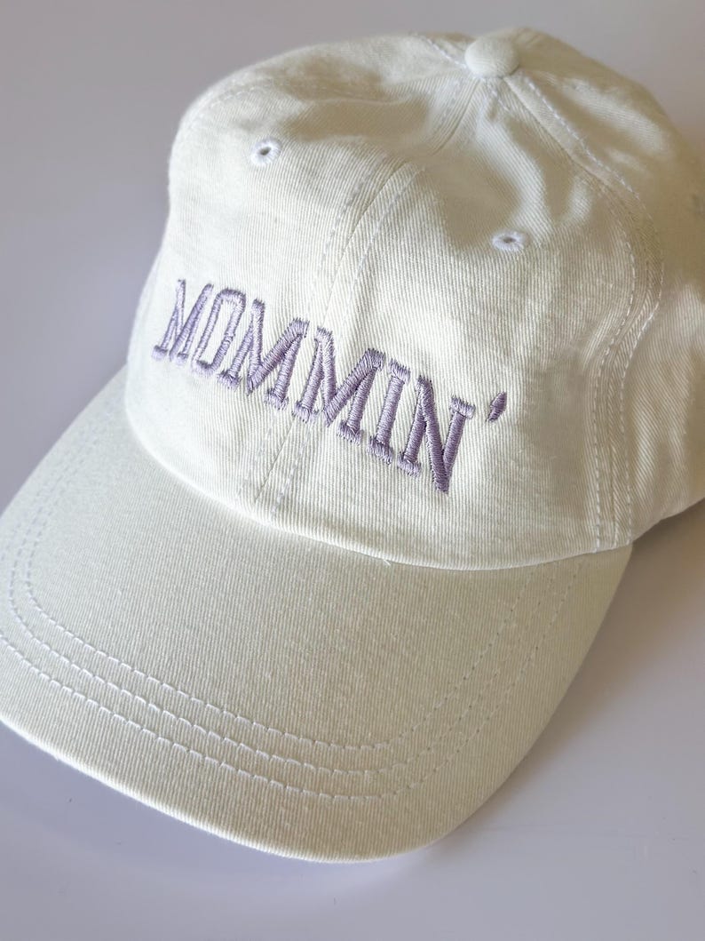 May include: A white baseball cap with the word "MOMMIN'" embroidered in purple on the front.