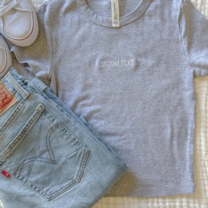 May include: A light grey t-shirt with the embroidered text "CUSTOM TEXT", alongside light blue denim jeans and white trainers. The t-shirt features a crew neck and short sleeves. The jeans are folded and have a red tag.