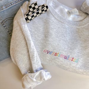 May include: A light grey heathered crewneck sweatshirt featuring the word overstimulated embroidered in a rainbow-coloured thread. A black and white chequered hair claw clip rests on the fabric, alongside a pair of light blue denim jeans.