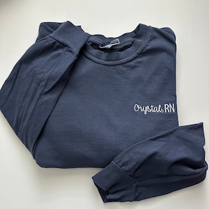 Op de afbeelding: Marineblauw shirt met lange mouwen met de geborduurde tekst "Crystal, RN" in het wit. Het shirt heeft een ronde hals en geribde manchetten. Het shirt is op een wit oppervlak gevouwen.