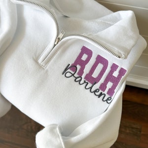 May include: A white quarter-zip sweatshirt featuring embroidered text on the left chest. The letters RDH are stitched in a purple block font, with the name Darlene embroidered underneath in a dark gray cursive script font.